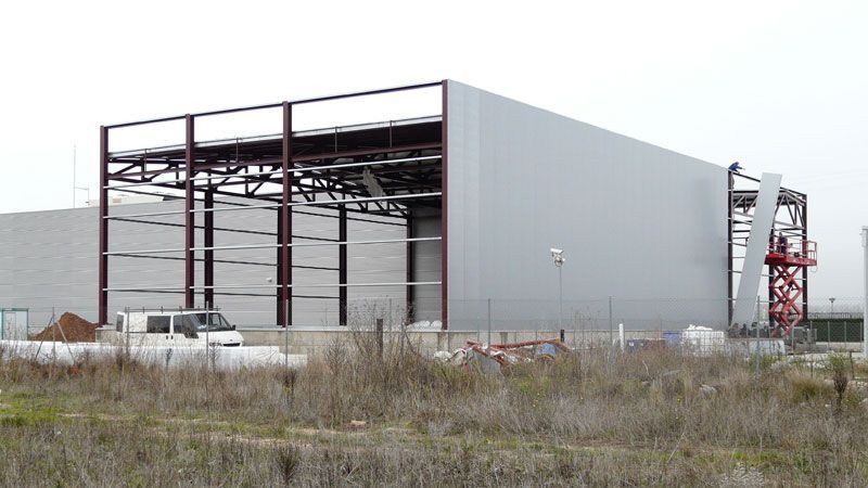 Gran edificio industrial en construcción con estructura de acero, paneles grises y césped crecido en la fachada.