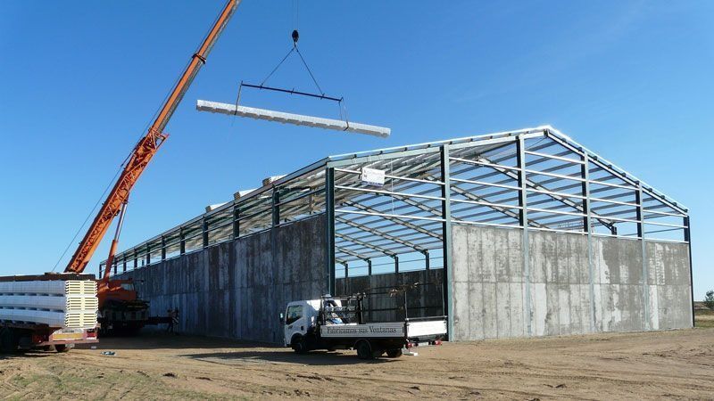 Obra en construcción con un almacén de estructura de acero, paredes de hormigón, una grúa levantando una viga y un camión cerca.