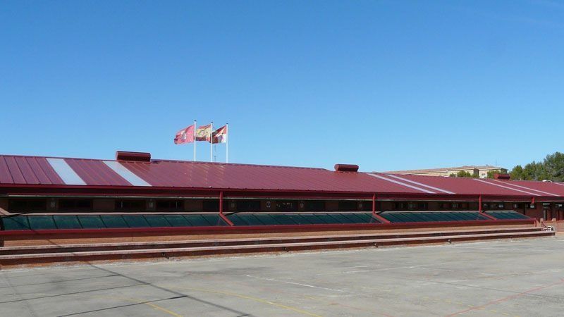 Edificio largo de techo rojo con mástiles de banderas bajo un cielo azul despejado.