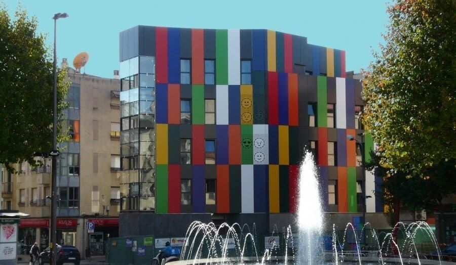 Edificio moderno y colorido junto a una fuente en una plaza de la ciudad bajo un cielo despejado.