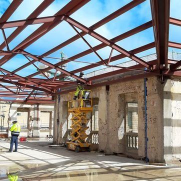 Trabajadores en una obra de construcción bajo una estructura de techo de acero rojo, junto a un edificio de hormigón.