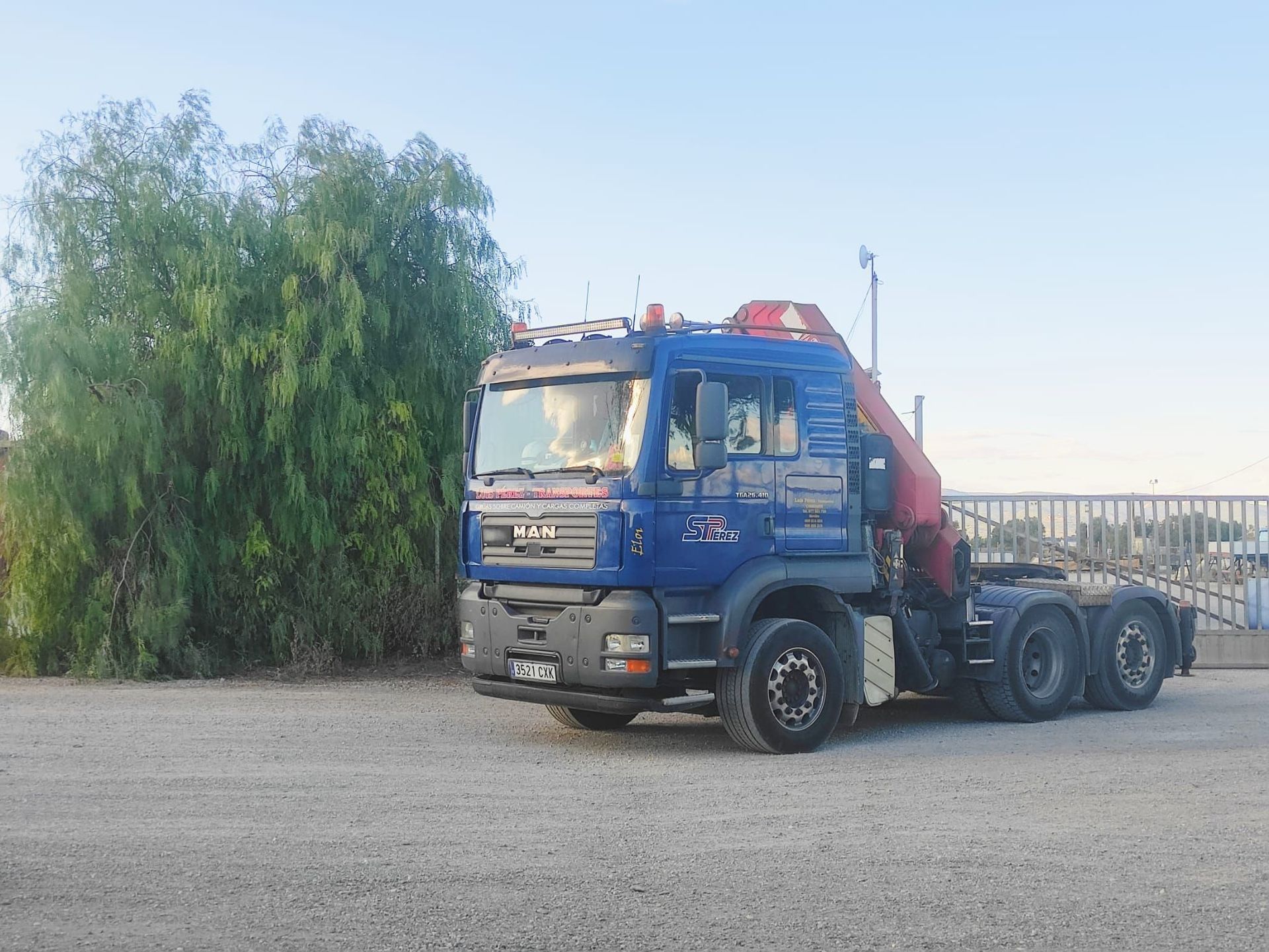 Un camión azul con una grúa roja en la parte trasera está estacionado en un lote de grava.