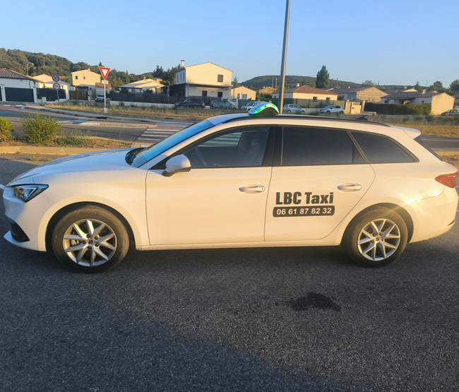 Taxi blanc avec l'inscription « LBC Taxi » et le numéro de téléphone sur le côté, stationné sur un parking.