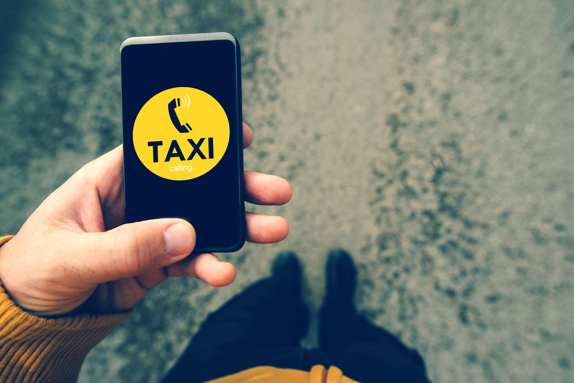 Une main tenant un smartphone avec une icône de taxi jaune, debout dans une rue, des jambes floues en dessous.