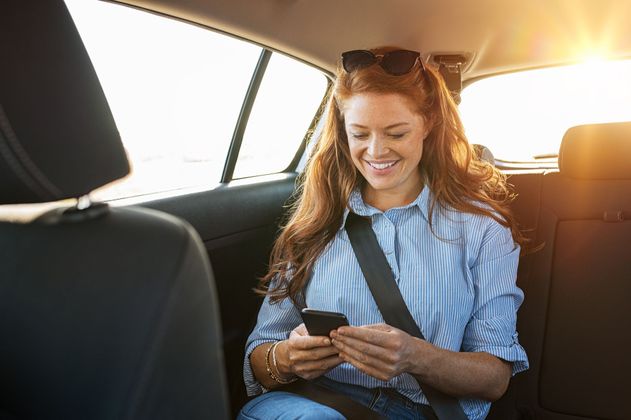 Une femme sourit à son téléphone sur la banquette arrière d'une voiture baignée de soleil.