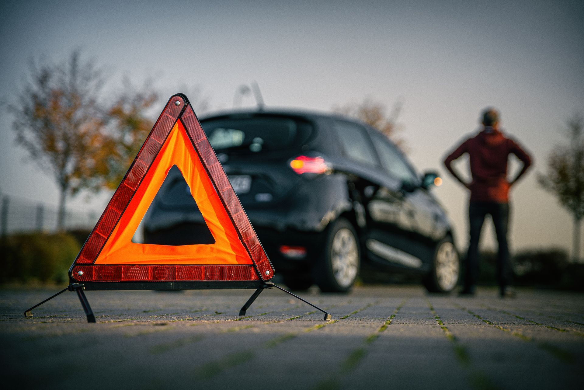 Triangle de signalisation réfléchissant en bord de route devant une voiture et une personne en panne sur la route