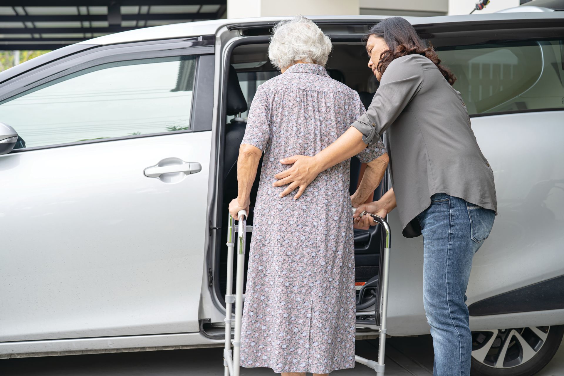 Une femme aide une personne âgée à monter dans une voiture argentée à l'aide d'un déambulateur, près de la porte arrière ouverte.