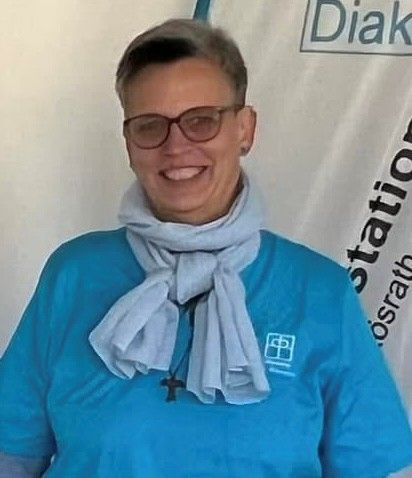 Eine lächelnde Person in einem blauen Hemd und einem hellen Schal, die eine Brille trägt, steht drinnen vor einem Wandschild.