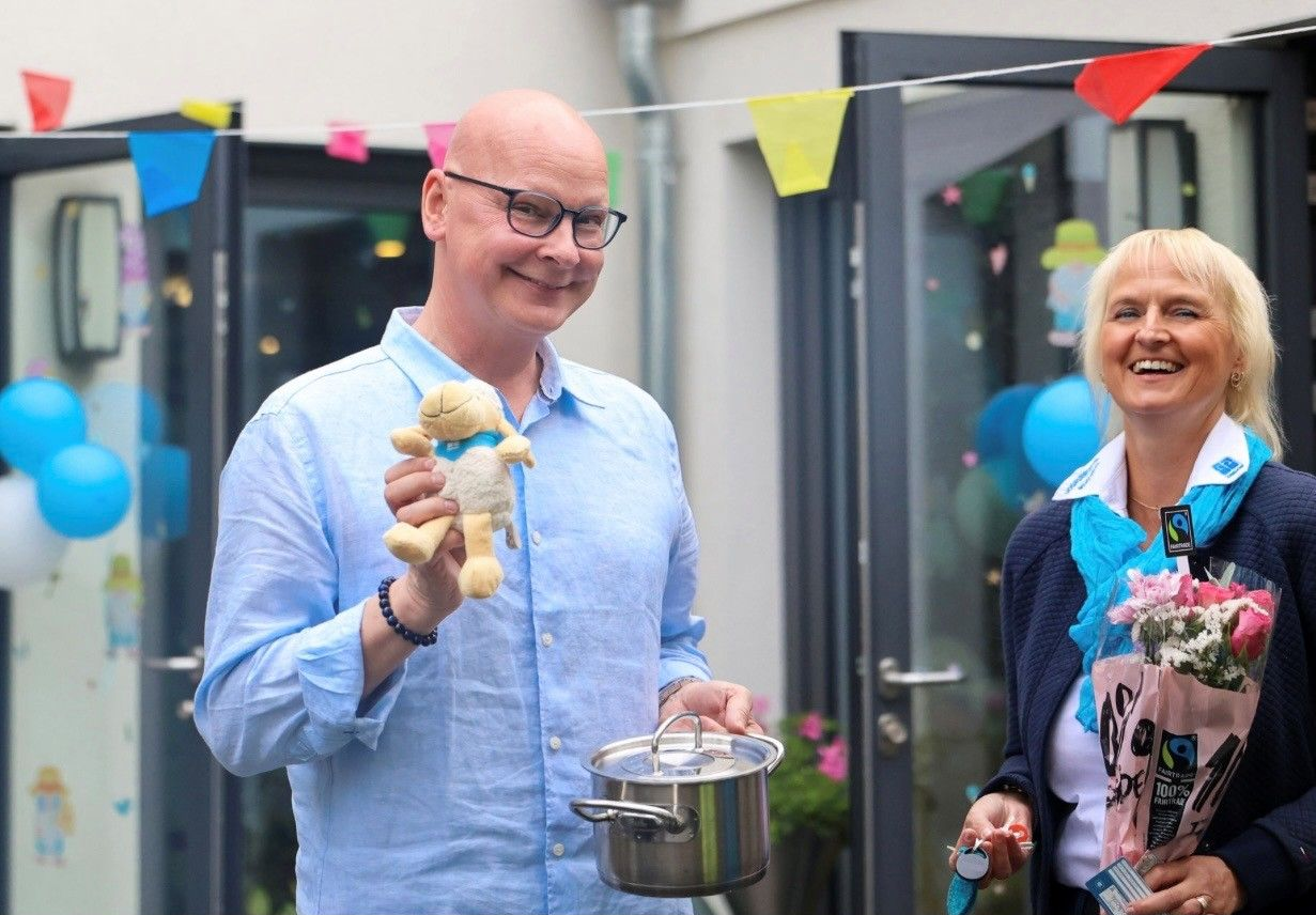 Zwei lächelnde Personen bei einer Feier im Freien mit Wimpelketten und Luftballons, die Leckereien und einen kleinen Topf in den Händen halten.