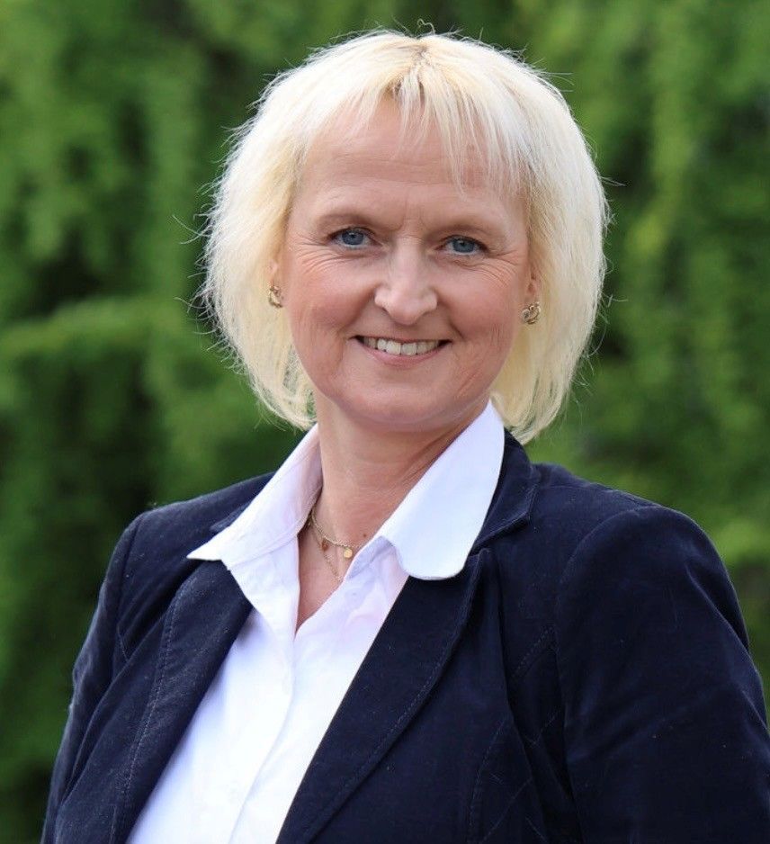 Eine lächelnde Person in einem weißen Hemd und einem dunklen Blazer steht im Freien; im Hintergrund ist verschwommenes Grün zu sehen.