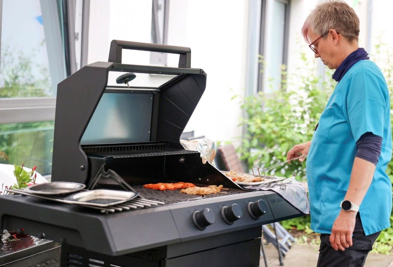 Eine Person grillt Essen auf einem schwarzen Grill in einer hellen Terrassenumgebung.