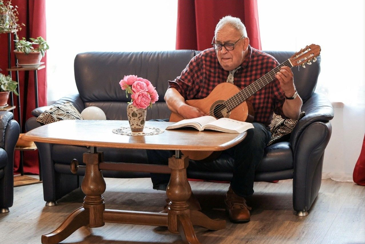 Ein Mann spielt Akustikgitarre auf einem Sofa im Wohnzimmer neben einem Couchtisch mit Blumen und Noten.
