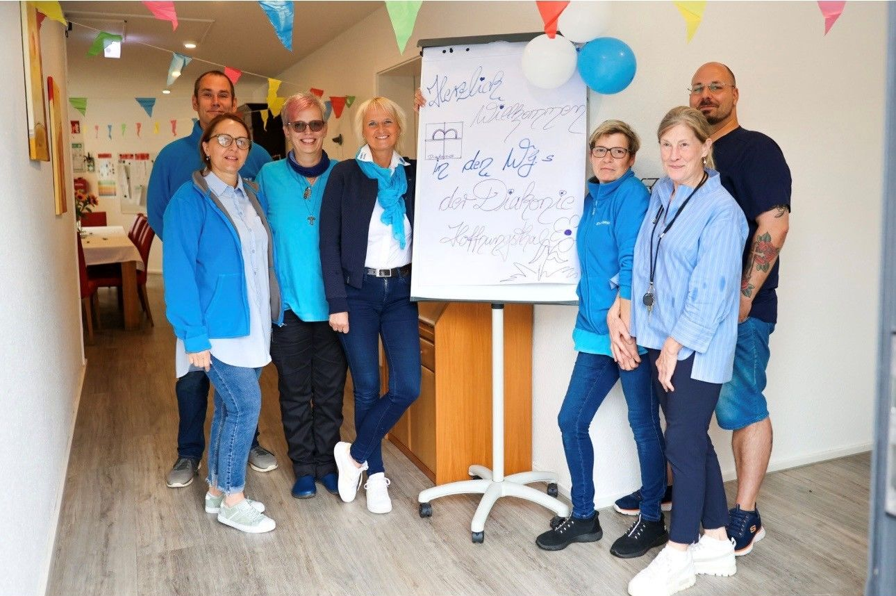 Eine Gruppe von Menschen posiert neben einer Tafel bei einer festlich geschmückten Feier in einem Innenraum; darüber hängen Luftballons und Wimpelketten.