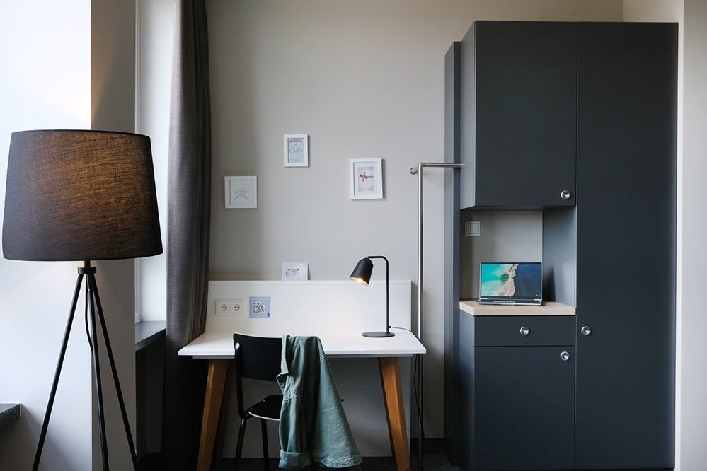 Ein Zimmer mit Schreibtisch, Stuhl, Lampe und Kleiderschrank.