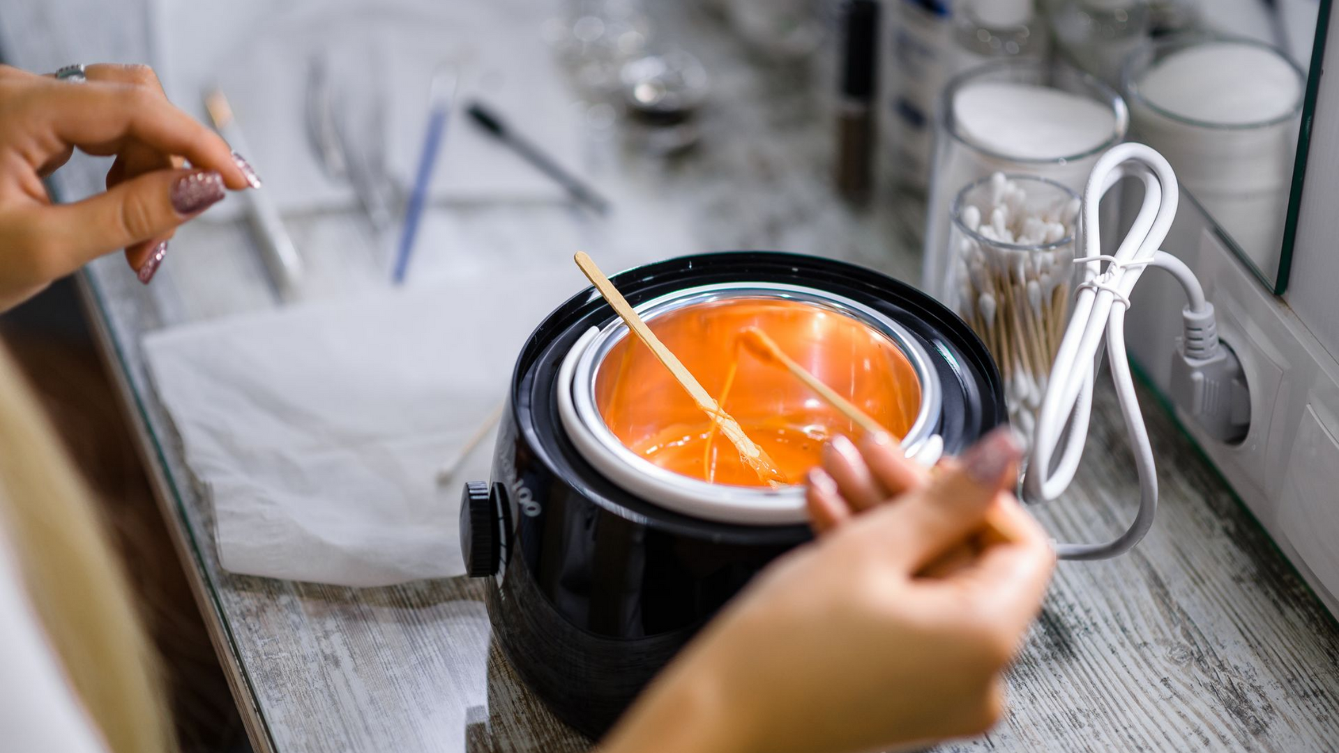 Une personne se prépare à s'épiler, en utilisant un chauffe-cire avec de la cire orange et des applicateurs en bois.
