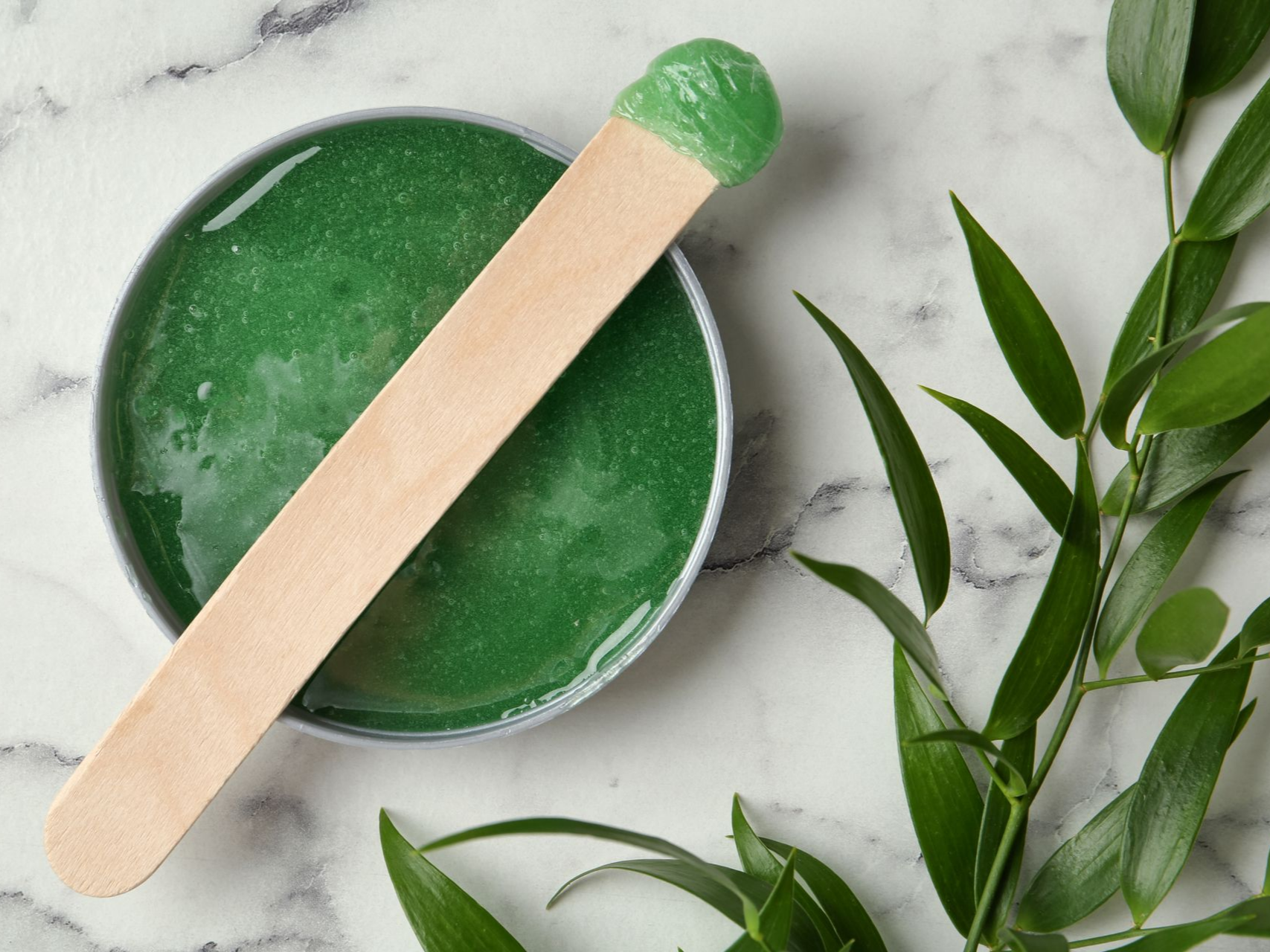 Cire dépilatoire verte dans un récipient en argent avec un applicateur en bois, feuilles sur le côté.