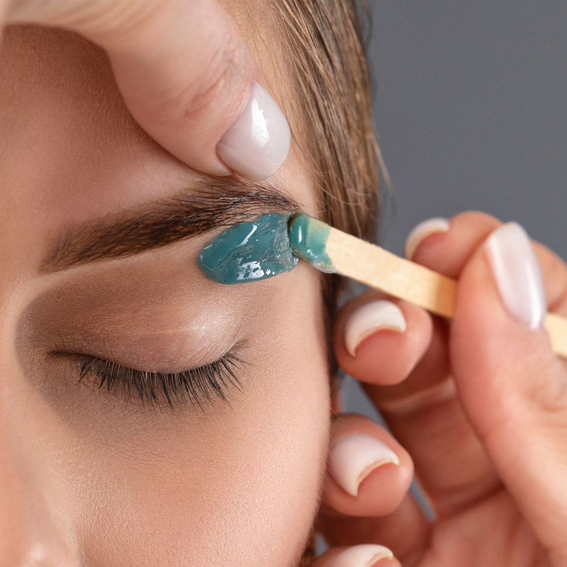 Application de cire sur les sourcils à l'aide d'un bâtonnet en bois ; œil fermé, expression neutre.