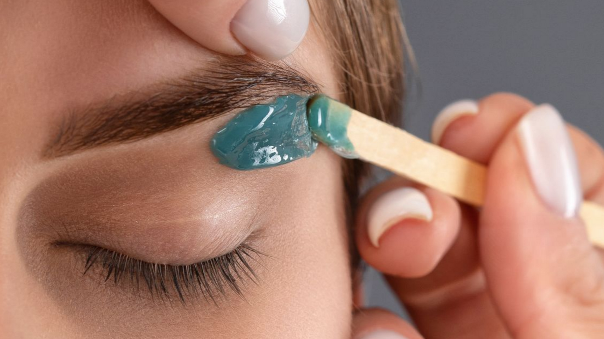 Épilation des sourcils à la cire bleue sur œil fermé, à l'aide d'un applicateur en bois.