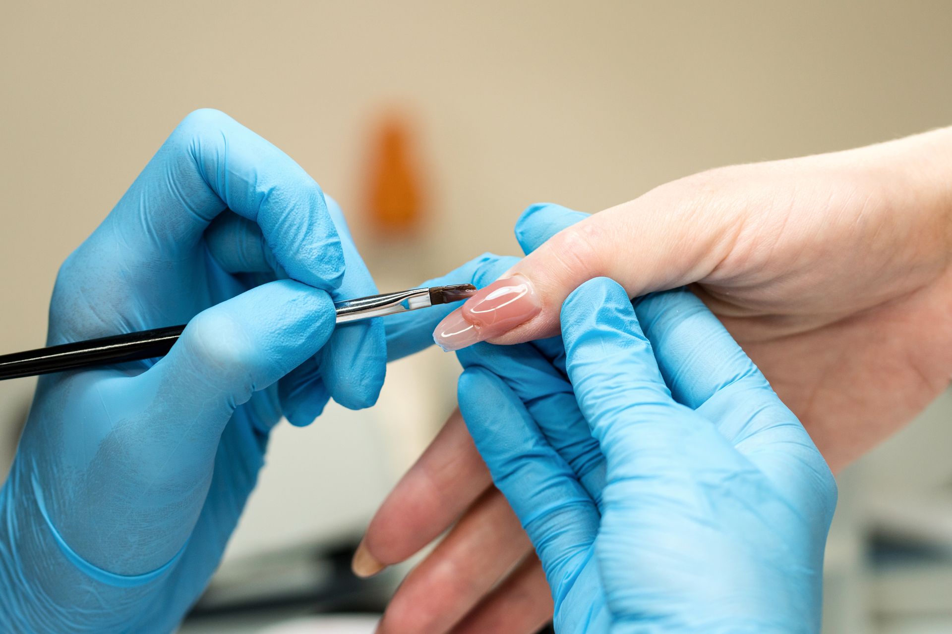 Des mains gantées de bleu appliquent du vernis à ongles transparent sur un ongle.