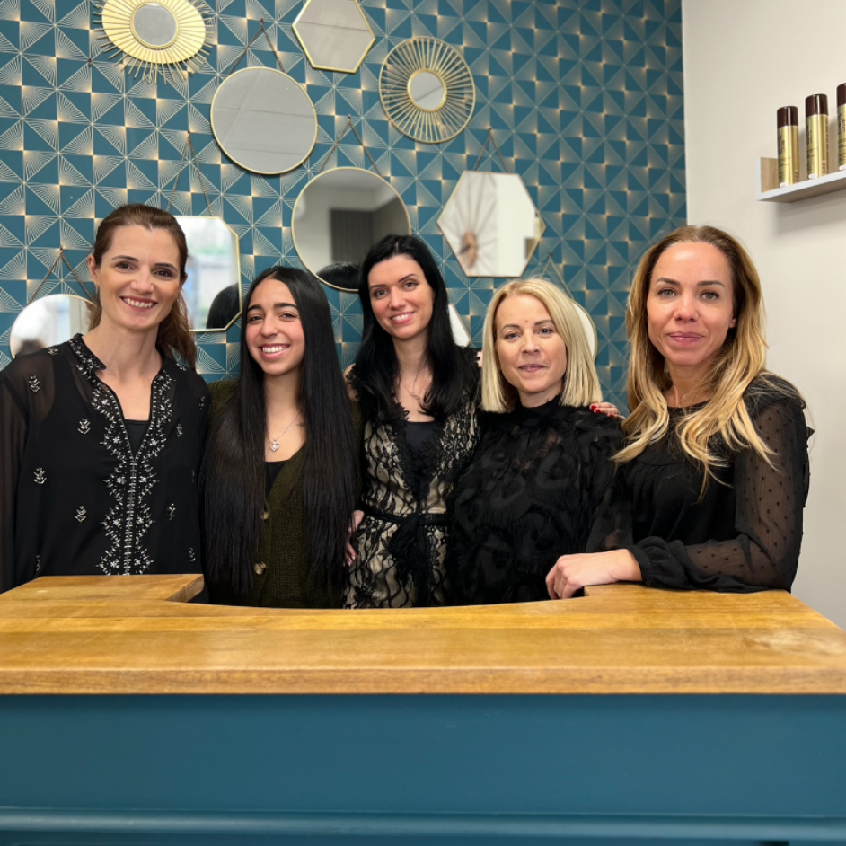 Cinq personnes dans un salon. Elles sourient derrière un comptoir en bois, devant un papier peint bleu et des miroirs.