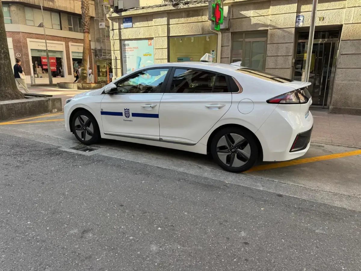 Un sedán blanco estacionado en una calle de la ciudad junto a un bordillo amarillo, con franjas azules en el lateral.
