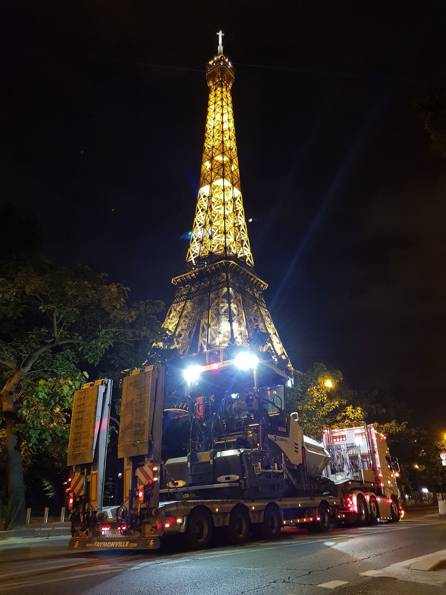 Poids lourd de dos transportant un véhicule de chantier avec vue sur la tour Eiffel, de nuit