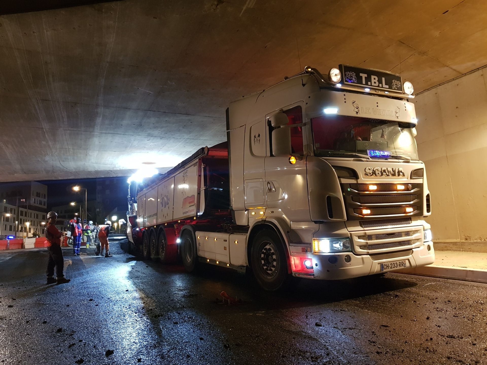 Plusieurs personnes autour d'un camion-benne sous un grand pont en ville