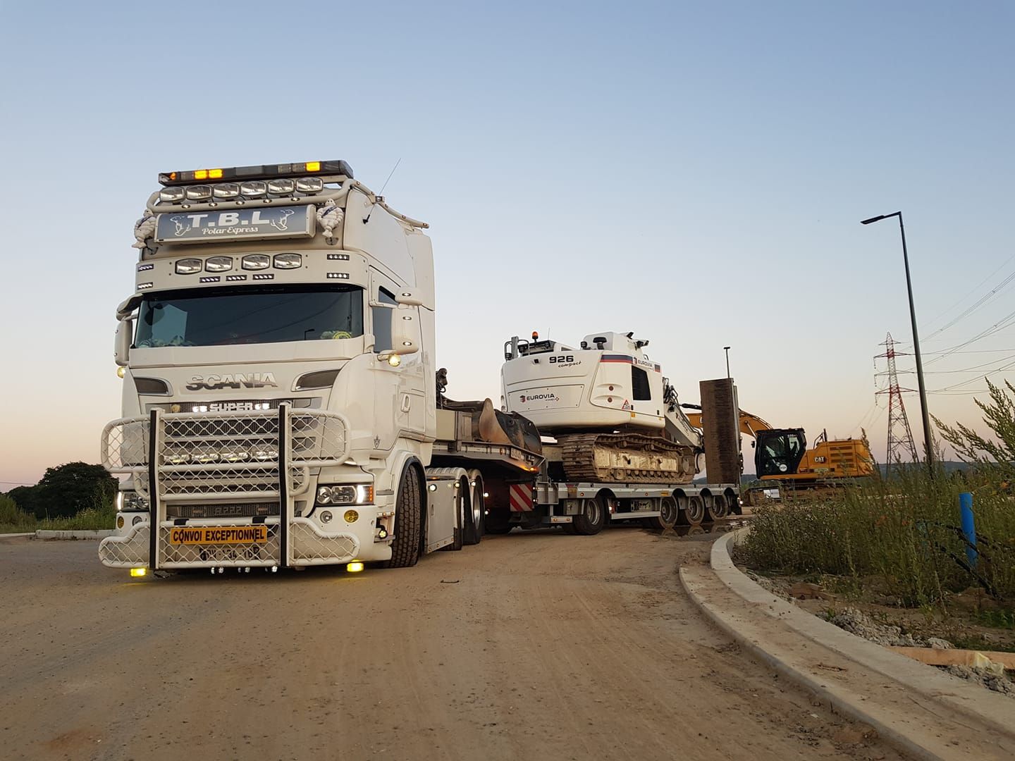Poids lourd blanc de chez TBL transportant un véhicule de chantier sur chenille