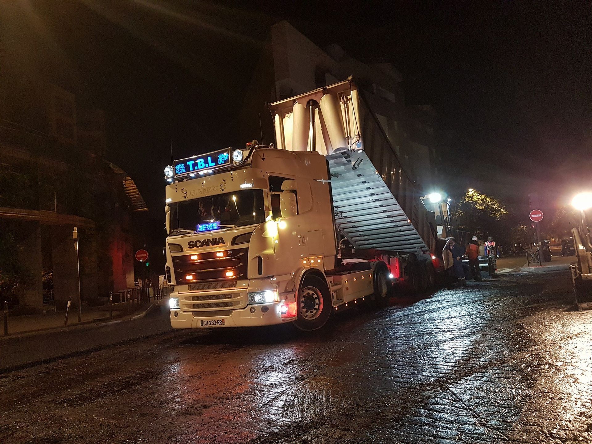 Camion-benne déversant son chargement d'enrobage sur une route en pleine ville, de nuit