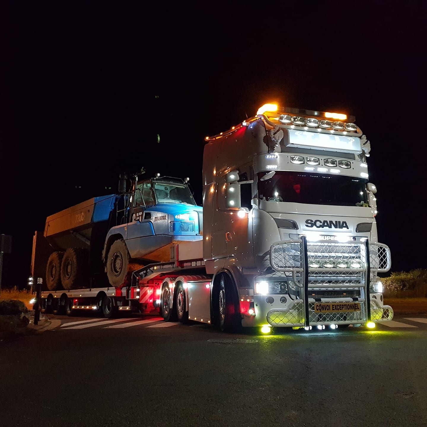 Camion blanc transportant un très gros véhicule de chantier bleu, de nuit