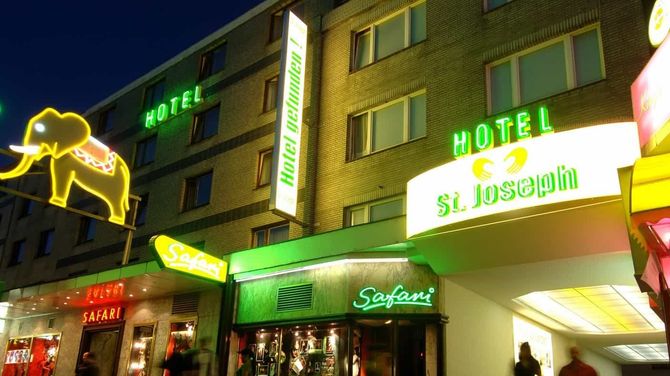 Der Eingang zum Hotel St. Joseph bei Nacht, mit einem Neon-Elefanten und grünen Schildern, und vorbeigehenden Menschen.