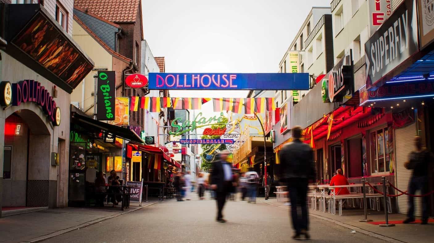 Straßenszene mit Leuchtreklamen, Geschäften und verschwommenen Fußgängern unter einem „DOLLHOUSE“-Schild.
