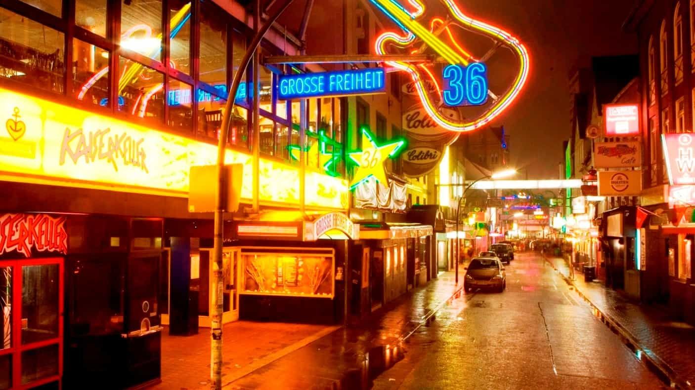 Nachts eine neonbeleuchtete Straße; mehrere Bars und Schilder erhellen die nasse Fahrbahn; ein gitarrenförmiges Schild.