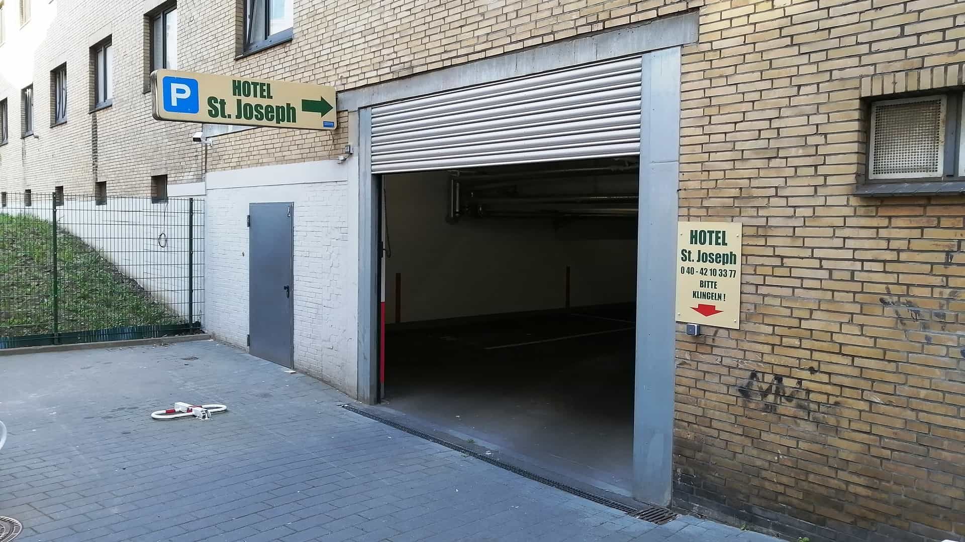Garageneinfahrt mit teilweise geöffnetem Metallrollladen. Ein Schild weist Richtung „St. Joseph“. Backsteinmauer.