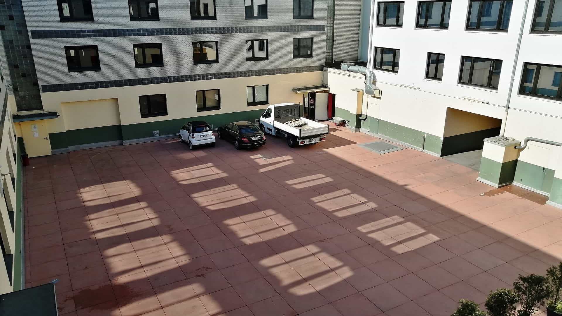 Autos parkten in einem Innenhof, die Schatten der Gebäude fielen auf die roten Fliesen.