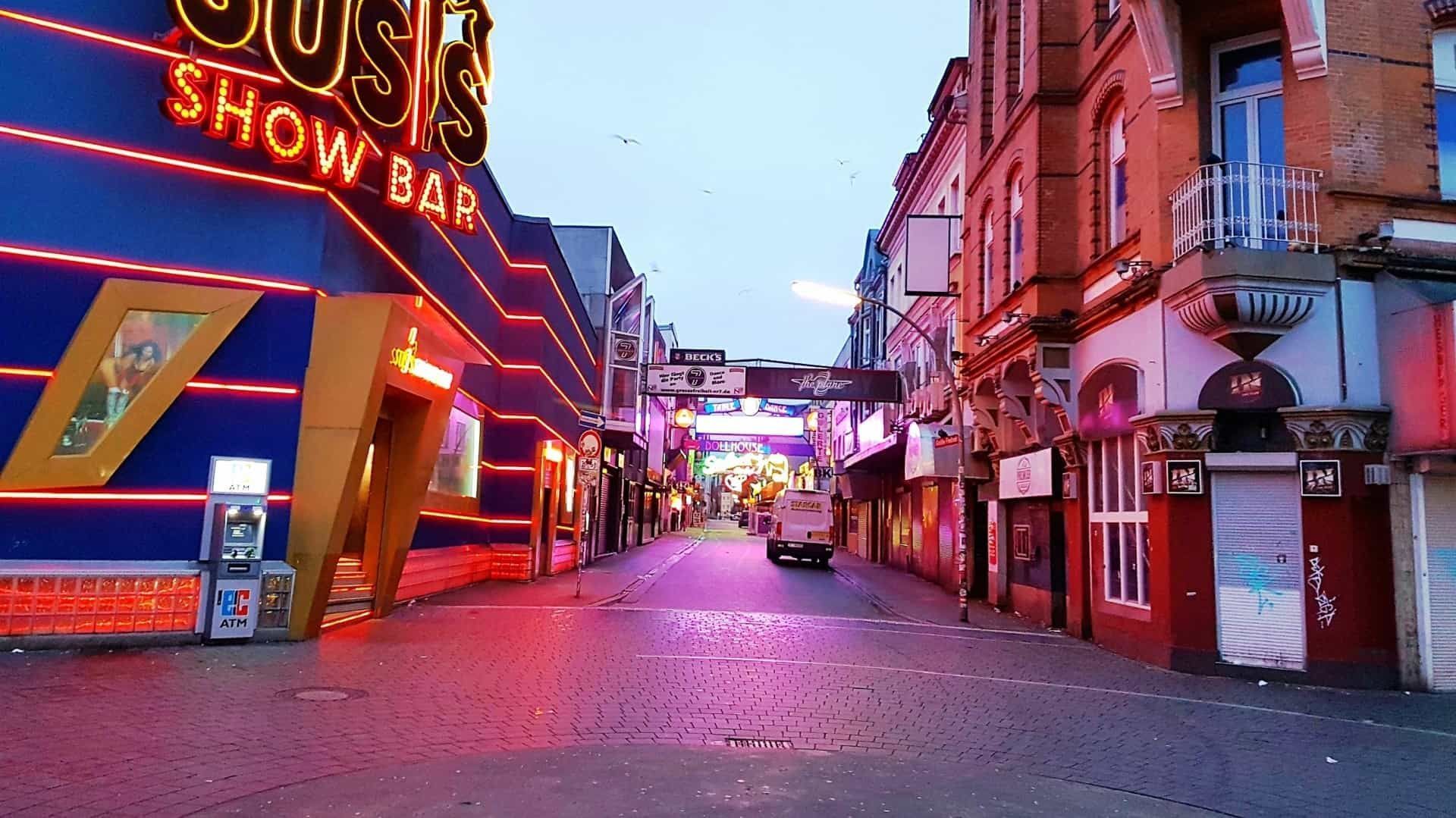 Straßenansicht mit Leuchtreklamen; links eine Showbar, Kopfsteinpflasterstraße, Gebäude auf beiden Seiten.