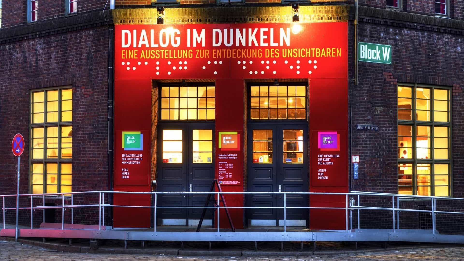 Roter Eingang des Museums „Dialog im Dunkeln“, Backsteingebäude, beleuchtete Schilder, Türen und ein Metallgeländer.