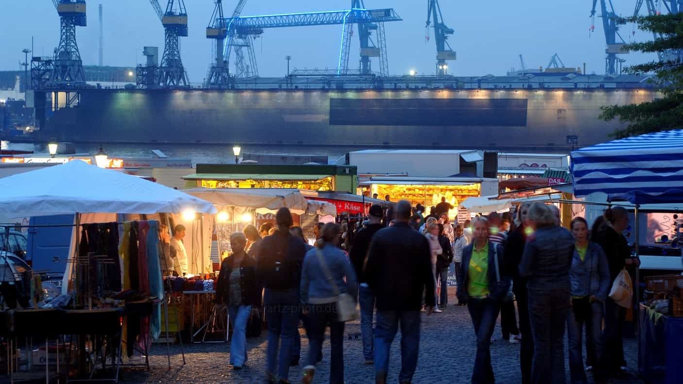 Menschen auf einem Abendmarkt vor der Kulisse einer Werft; Lichter beleuchten die Verkaufsstände und die Menschenmenge.