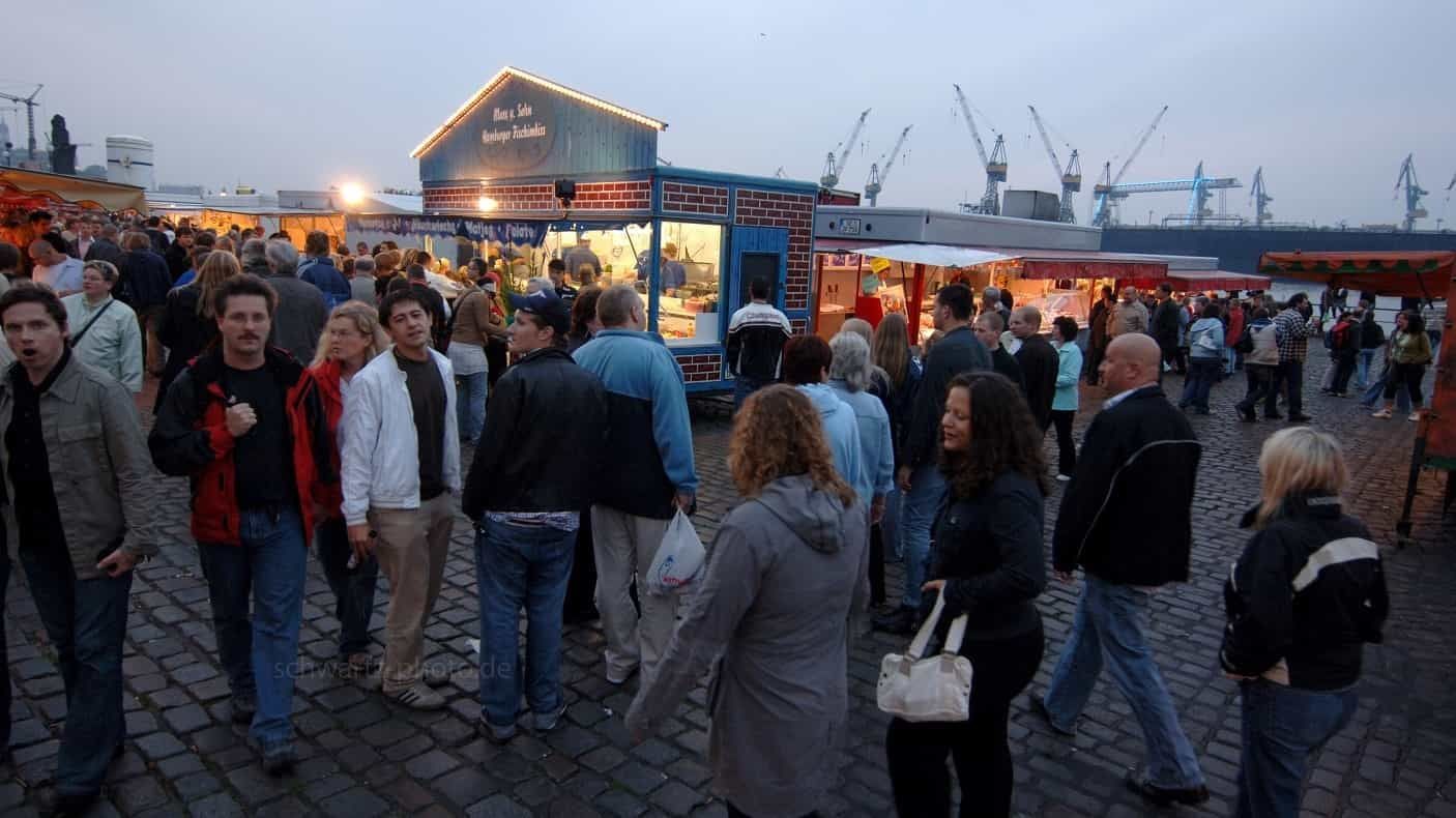 Menschen auf einem Freiluftmarkt mit Essensständen in der Abenddämmerung. Kräne und Wasser im Hintergrund.