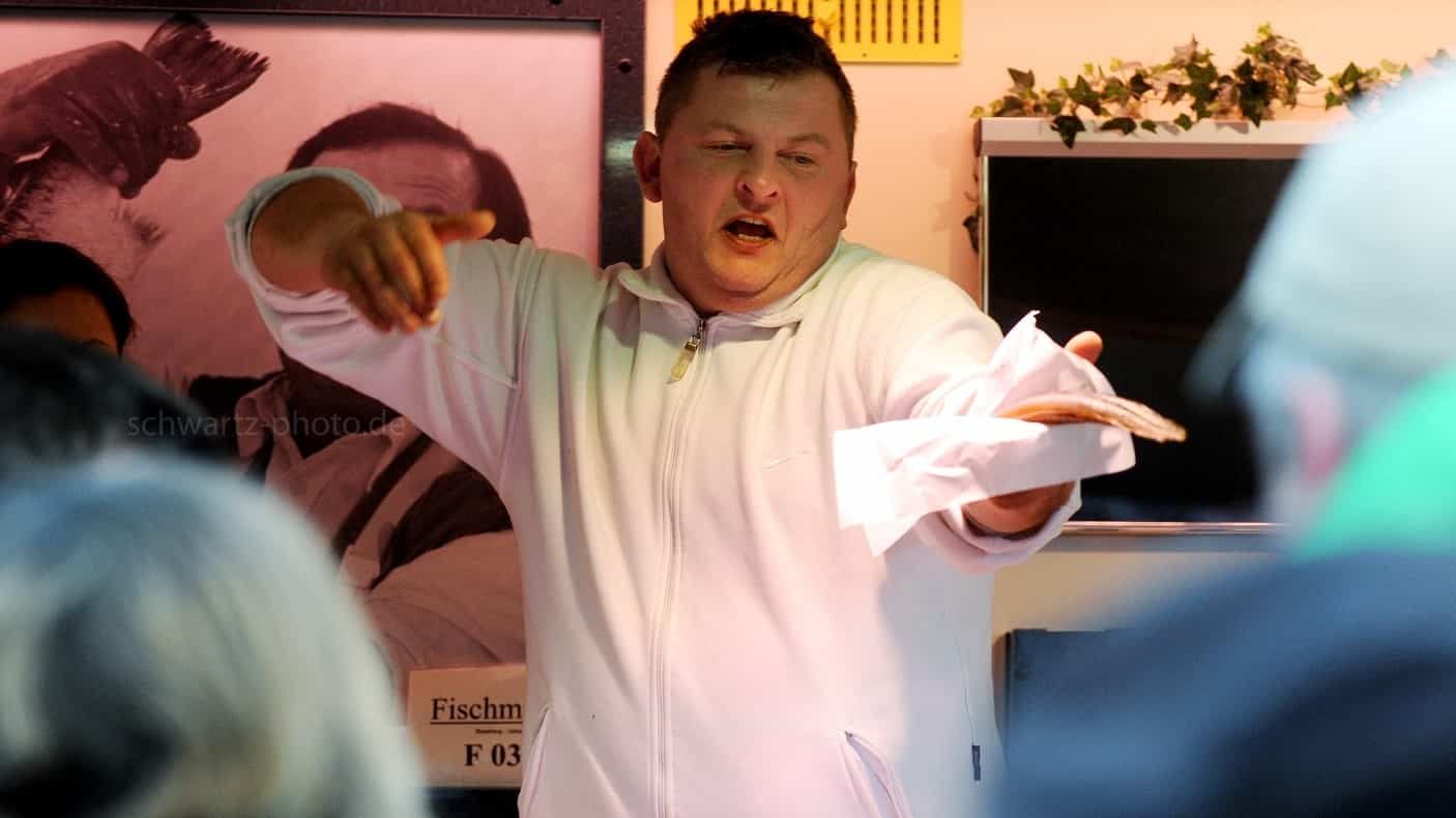 Ein Mann im weißen Kittel gestikuliert, hält ein Blatt Papier in der Hand und spricht zu einer Gruppe.