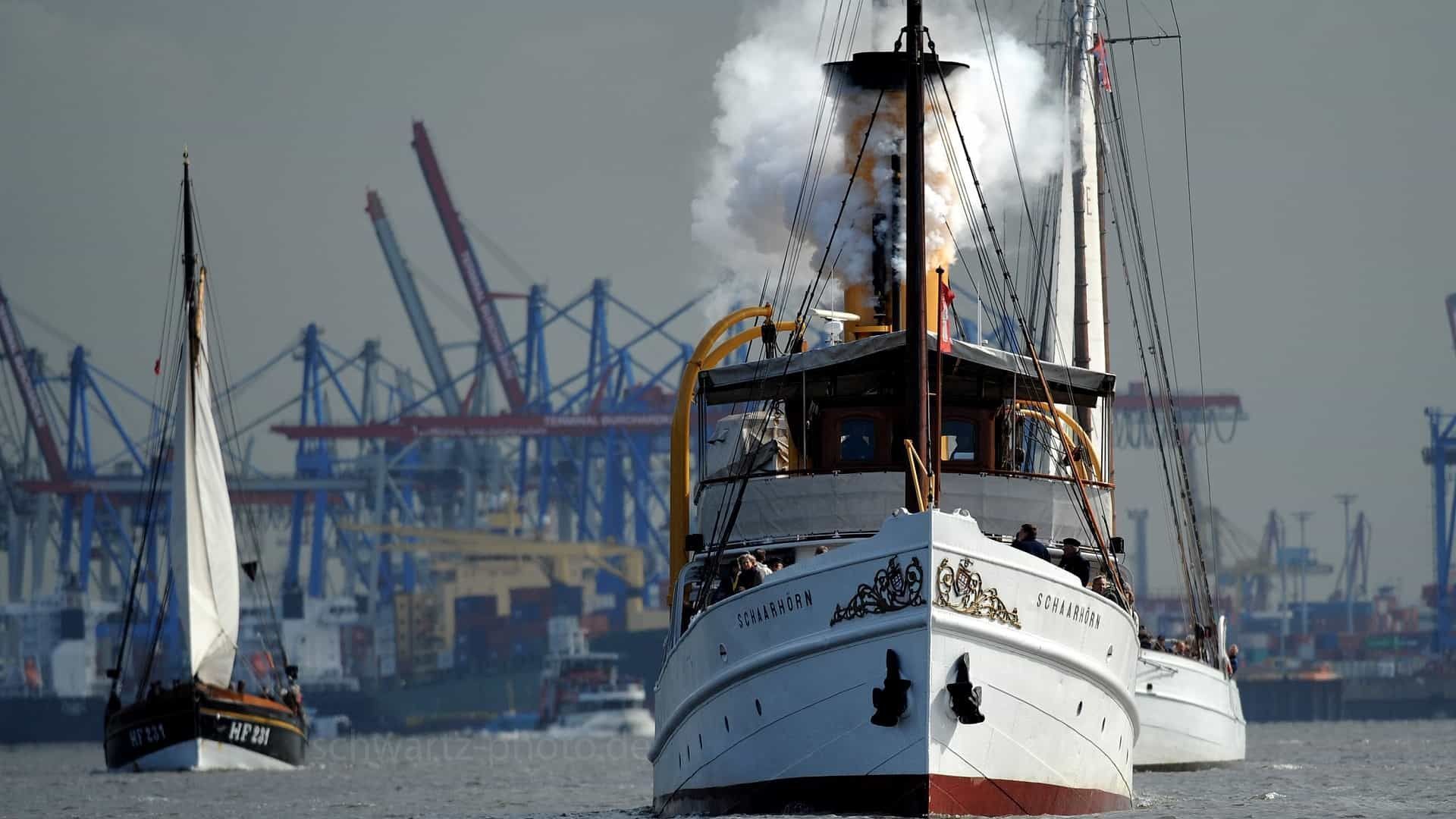 Historisches Dampfschiff mit Rauchentwicklung, das an anderen Booten in einem Hafen vorbeifährt, Kräne im Hintergrund.