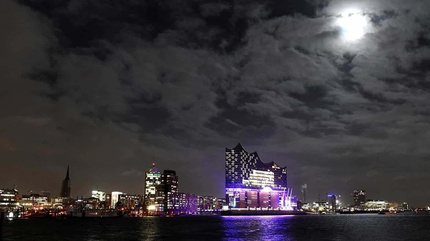 Nachtansicht einer Stadtsilhouette mit hellem Mond, Wolken und beleuchteten Gebäuden, die sich im Wasser spiegeln.