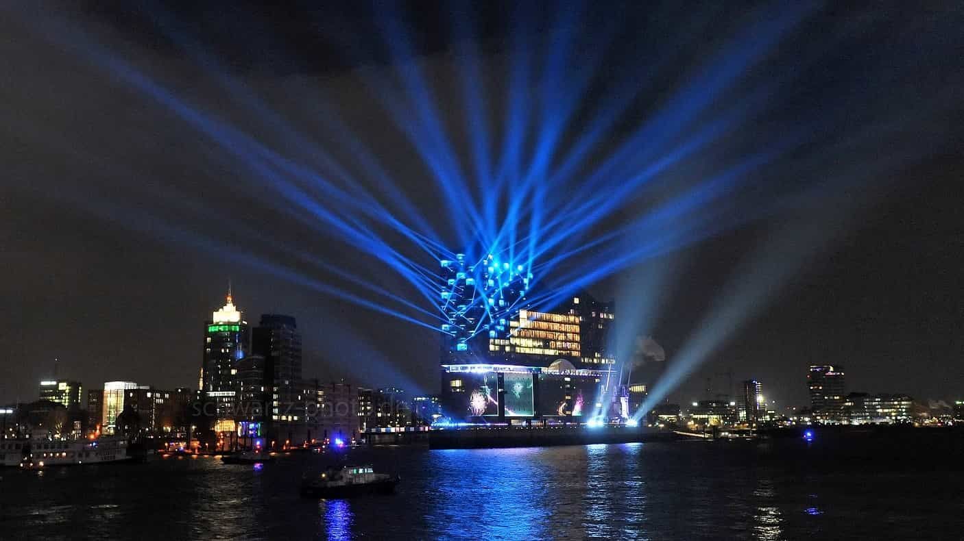 Nachtansicht einer Stadtlandschaft mit blauem Lichtspiel, das von einem Gebäude über dem Wasser ausgeht.