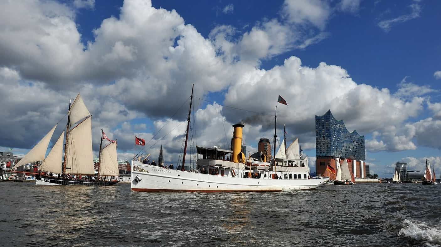 Boote fahren auf einem Fluss, im Hintergrund die Elbphilharmonie unter blauem Himmel mit Wolken.