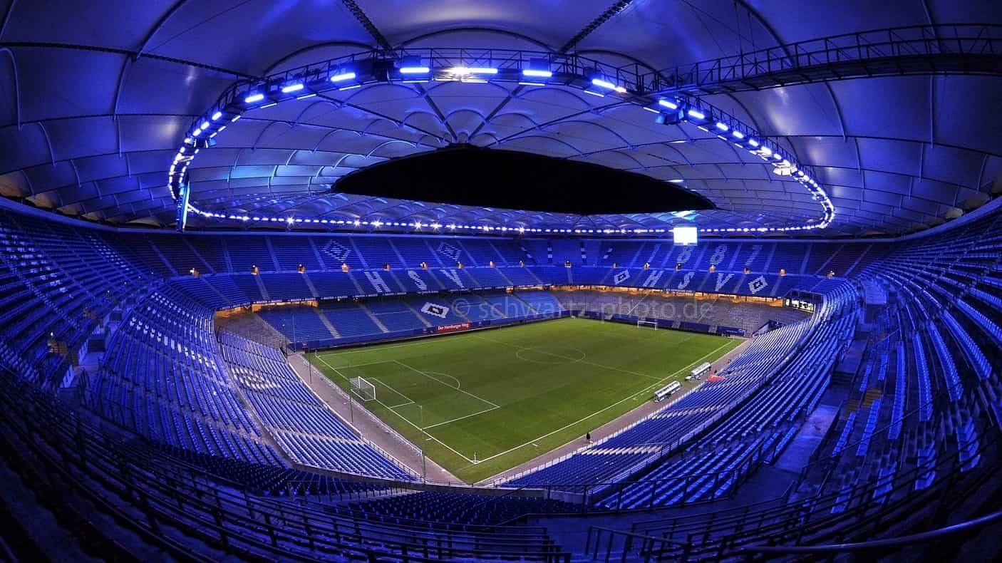 Blau beleuchtetes Stadioninterieur mit grünem Spielfeld, leeren blauen Sitzen und offenem Dach.