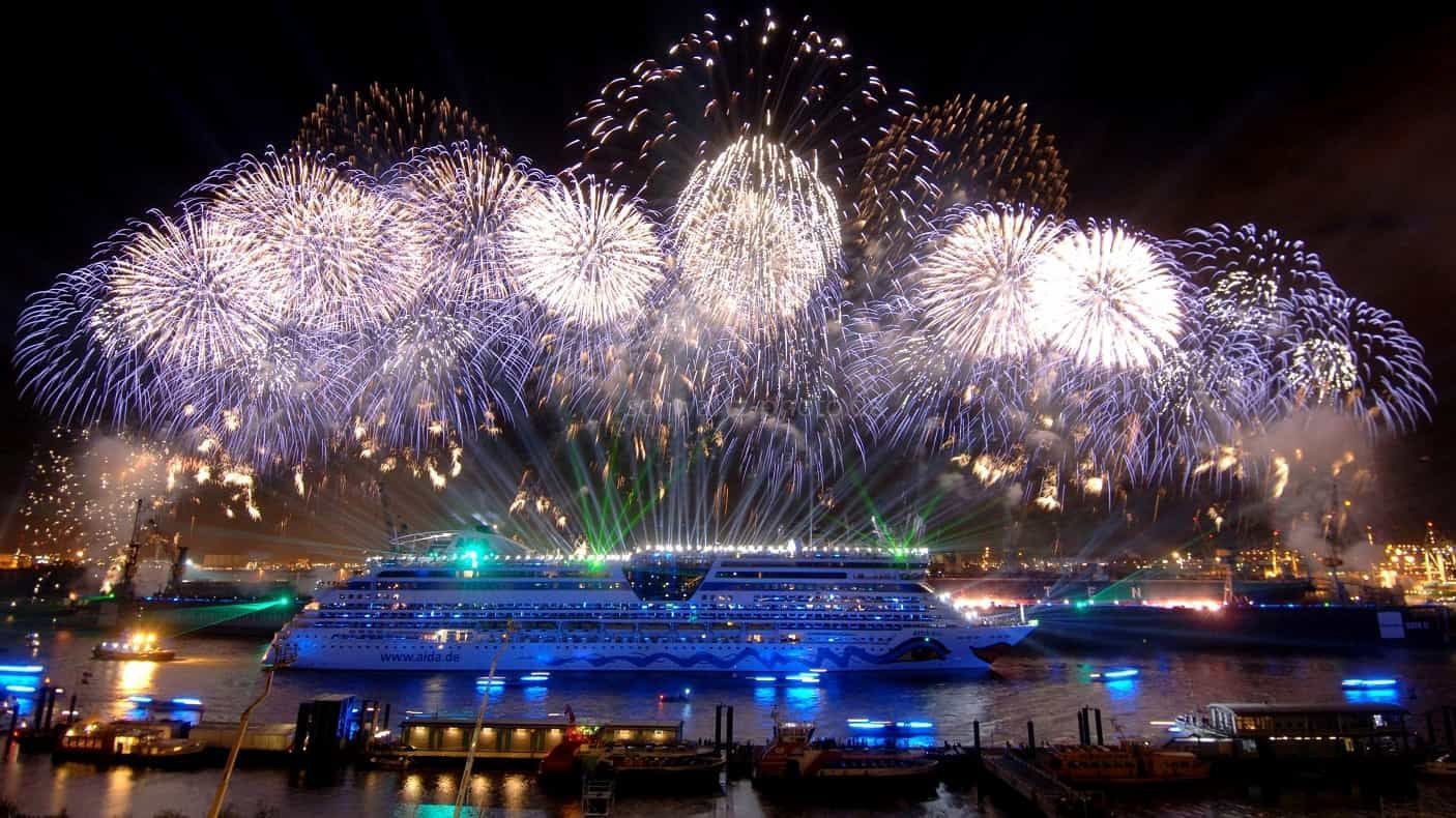 Feuerwerkskörper explodieren nachts über einem beleuchteten Kreuzfahrtschiff auf dem Wasser.