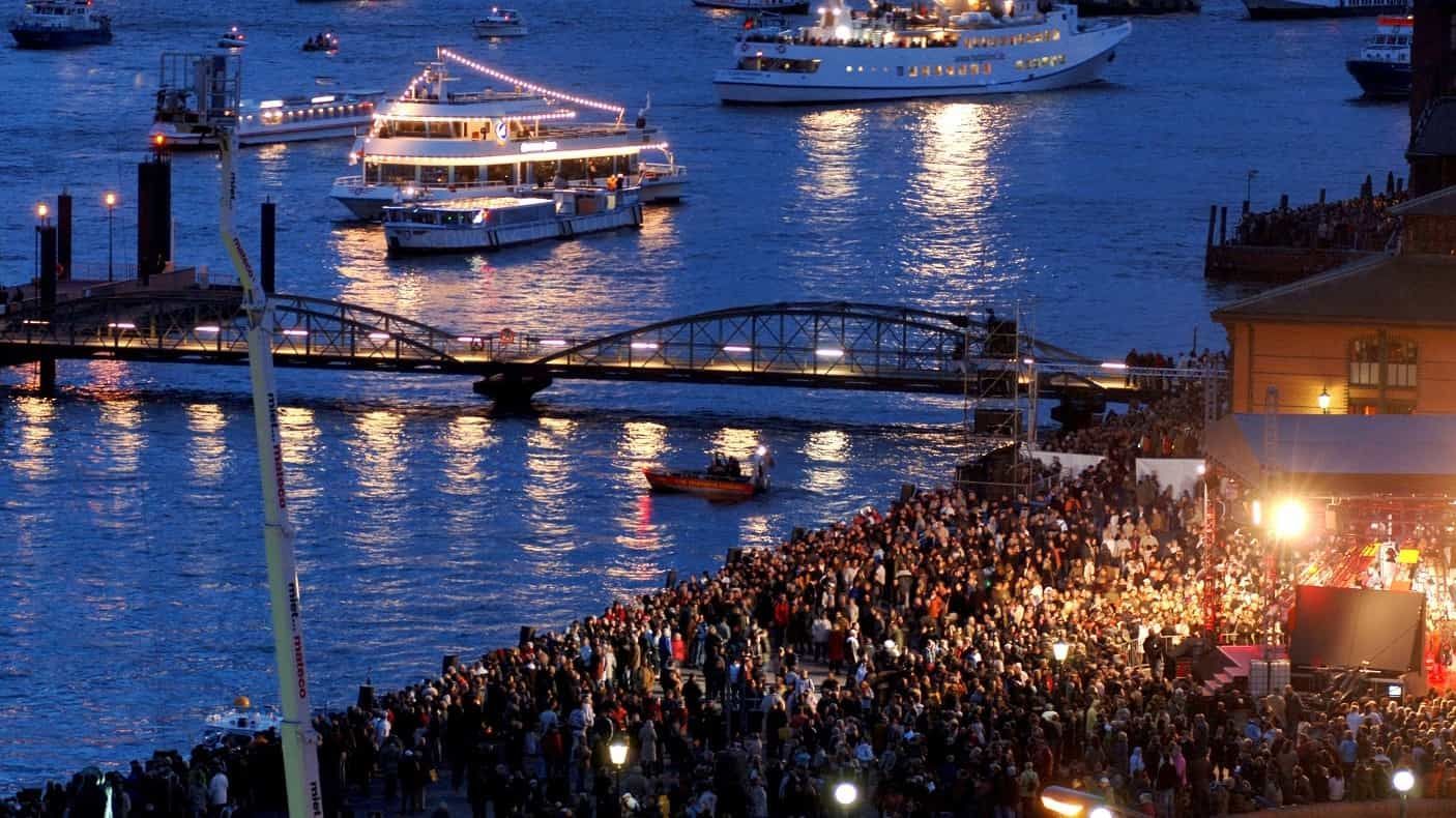 Große Menschenmenge bei einer Veranstaltung am Wasser; Boote mit Lichtern auf dem Wasser und eine Brücke.