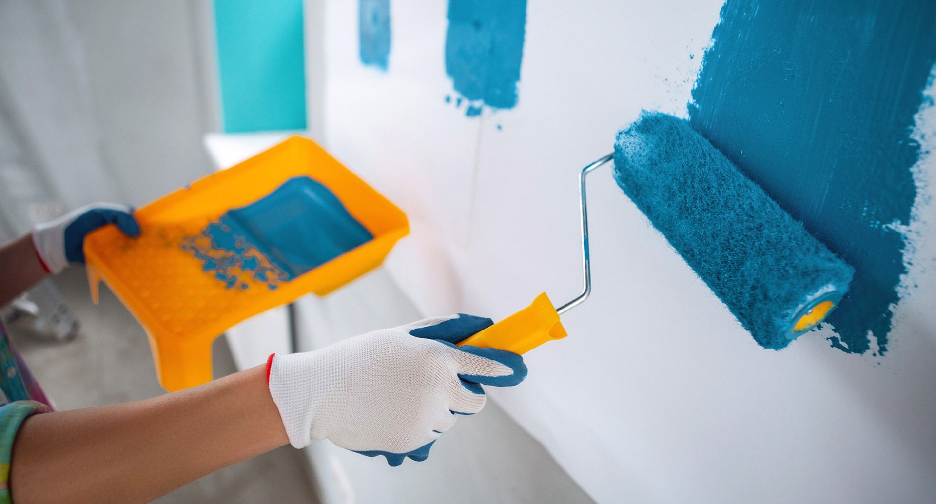 Una persona que lleva guantes utiliza un rodillo para pintar una pared con pintura color turquesa.