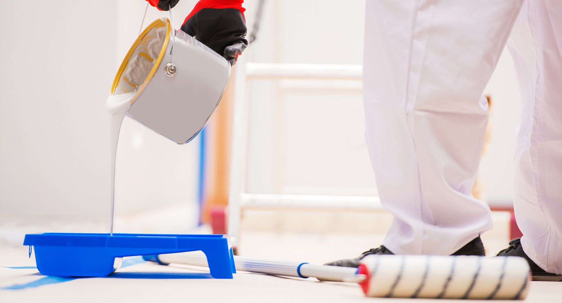 Una persona vestida con ropa de trabajo blanca vierte pintura blanca de un cubo en una bandeja de pintura azul.