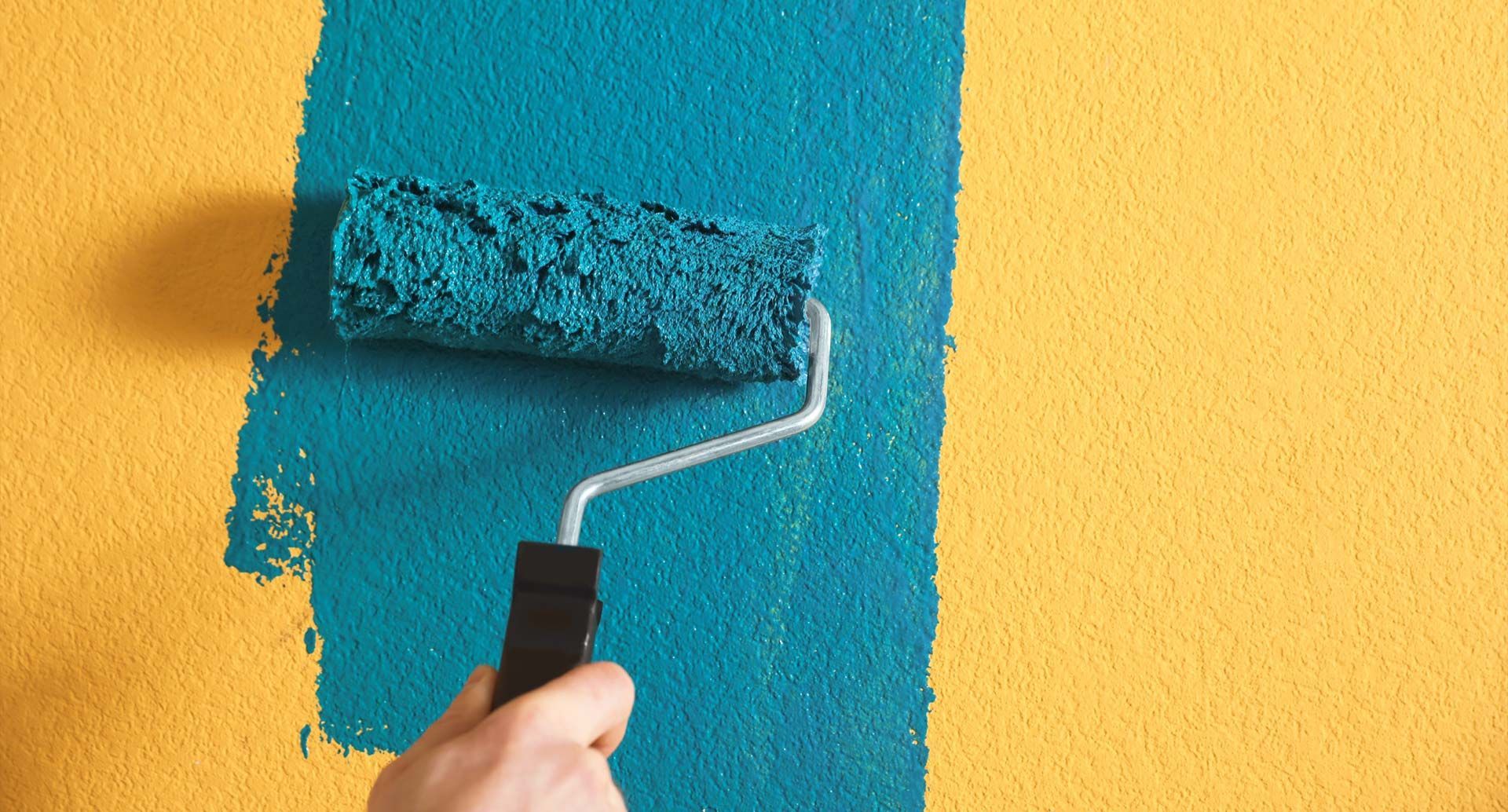 Una mano utiliza un rodillo para aplicar pintura color turquesa sobre una pared amarilla texturizada.